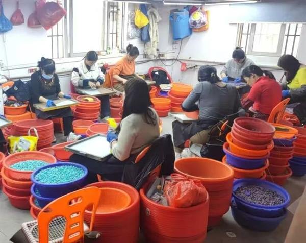 Workers sorting coloured beads by hand at benches, with stacked trays